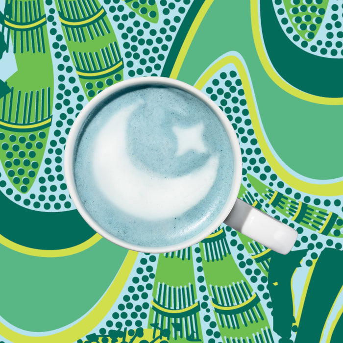 The Sleeptime Organic Superfood Latte by Kuli Kuli Foods, a blue latte infused with calming chamomile, displays a moon and star in the foam and rests on a bold, abstract green and yellow patterned background.