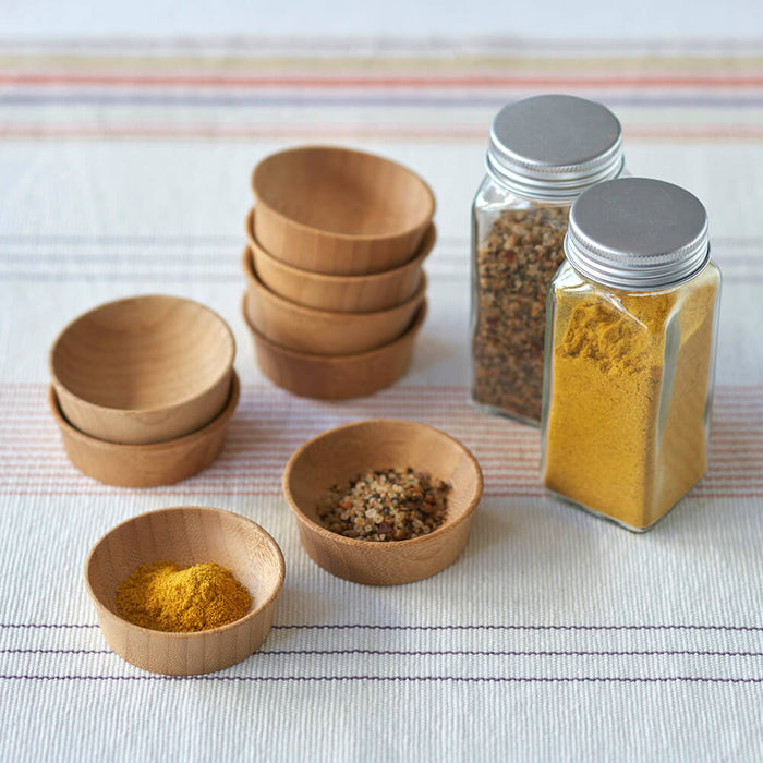 Small Bambu wooden condiment cups, similar to organic bamboo bowls, are filled with colorful spices like turmeric and mixed seasoning on a striped fabric. Nearby are clear jars with silver lids containing more spice mixtures, while empty wooden bowls lie waiting.