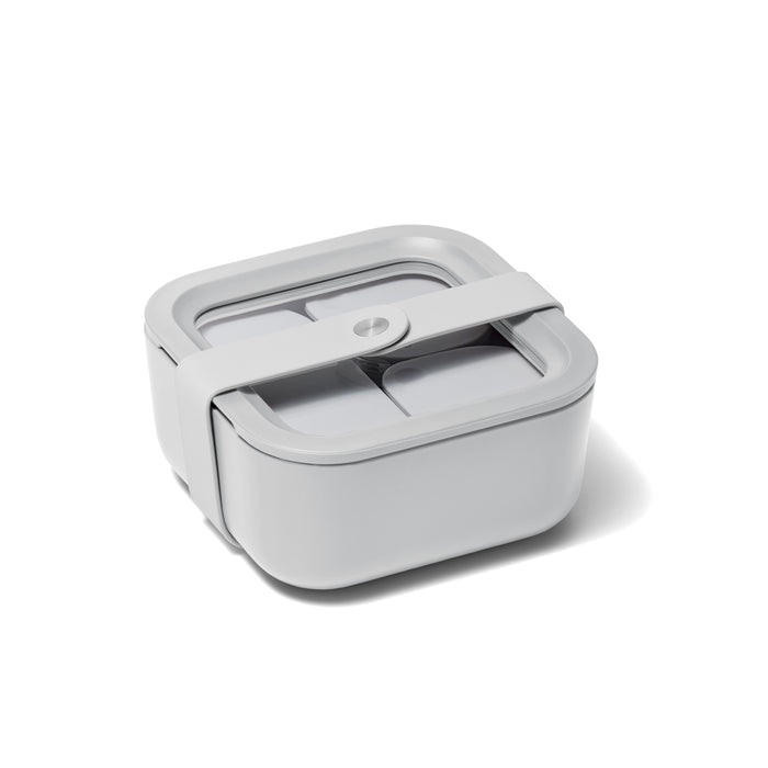 The Caraway Small Glass Food Container features a sleek, light gray square design with a white strap and a two-compartment lid, making it perfect for keeping meals organized and fresh.