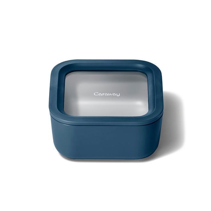 A Small Glass Food Container by Caraway, this square teal container features Air Release technology on its transparent lid with Caraway printed on top. The ceramic-coated glass has rounded edges and is shown on a plain white background.