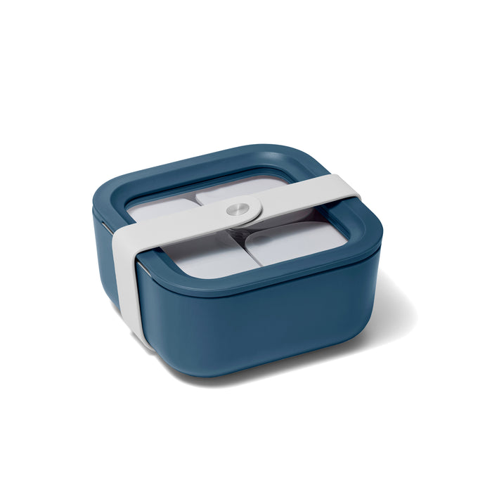 The Caraway Small Glass Food Container is a square, teal lunchbox featuring a transparent lid with four compartments. It has a white strap and uses ceramic-coated glass and Air Release technology to keep food fresh.