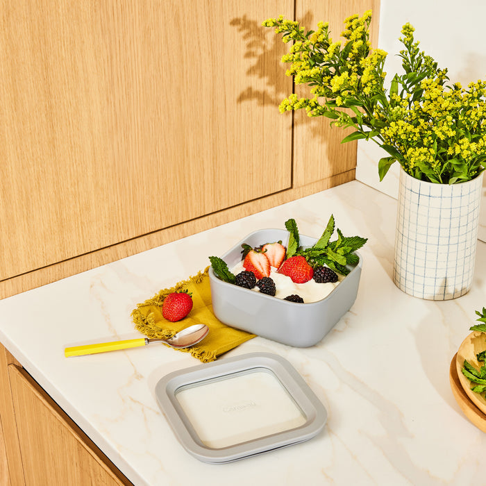 A small Caraway glass container, ceramic-coated and gray, holds a bowl of yogurt with strawberries, blackberries, and mint on a kitchen counter. A yellow-handled spoon lies on a matching cloth beside it, while yellow flowers in a vase stand near the wooden cabinet in the background.