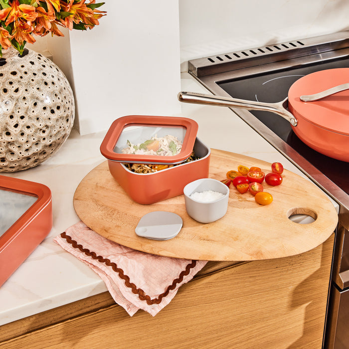 In a kitchen scene, a Caraway Small Glass Food Container with Air Release technology sits on a round wooden cutting board. Chopped tomatoes and a small white bowl are close by. An orange pot cooks on the stove, with a decorative vase filled with flowers in the background.