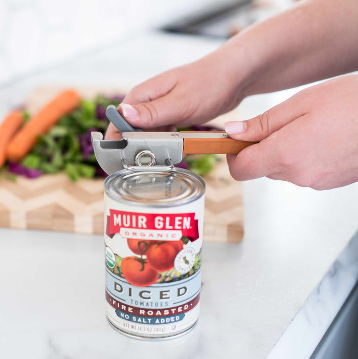 Hands skillfully using a Full Circle Smooth-Edge Can Opener to reveal the delightful goodness of Muir Glen Organic Diced Tomatoes, while carrots and greens await their turn on a wooden cutting board, with the safe smooth-edged lid ensuring worry-free handling in your cooking space.