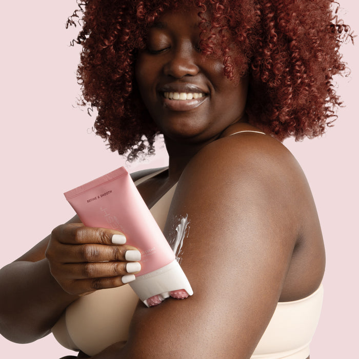 A person with curly hair, smiling in a beige top against a soft pink backdrop, applies VOESH New Yorks Smoothd Body Refining Roller Creme from a pink tube to their arm, enjoying the soothing moisture.