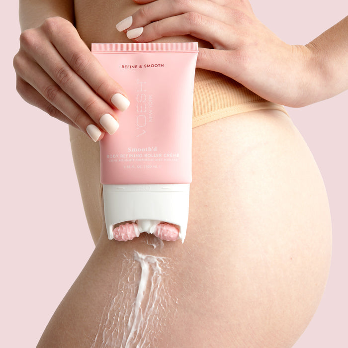 A person applies VOESH New Yorks Smoothd Body Refining Roller Creme, containing hyaluronic acid, onto their thigh. The cream glides from a pink tube with a roller applicator. Sporting neutral nails and a nude garment, they target cellulite for smoother skin.