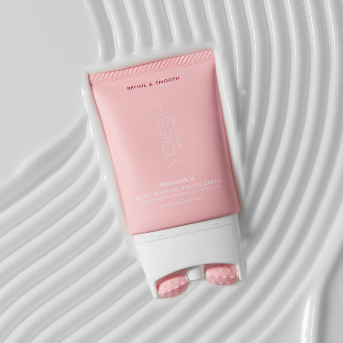 A pink tube of Smoothd Body Refining Roller Crème by VOESH New York, designed to fight crepey skin with integrated rollers, is set on a textured white surface. The chic packaging highlights the brand and product details.