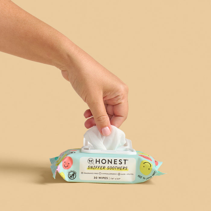 A hand pulls a wipe from a package of The Honest Company Sniffer Soothers, dermatologist-tested and scented with watermelon basil. The turquoise package has colorful fruit illustrations and contains 30 Gentle Soothing Nose and Face Wipes against a beige backdrop.
