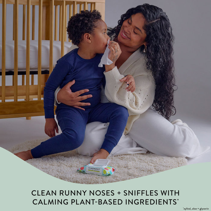 A woman lovingly wipes a young child's nose with a tissue. They are sitting on a fluffy rug in a cozy room with a crib nearby. A pack of tissues is on the floor, and text below reads, "Clean runny noses + sniffles with calming plant-based ingredients.