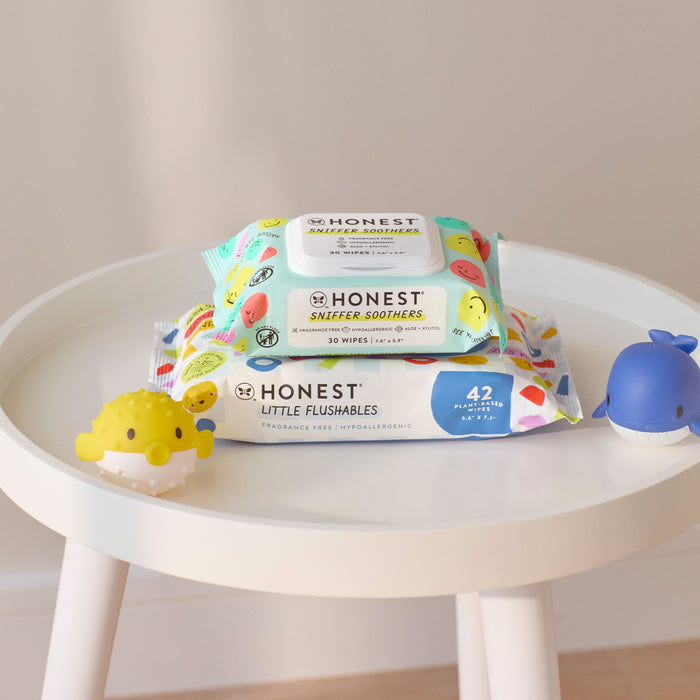 A small white table holds two packs of The Honest Company wipes with soothing ingredients. The top pack, Sniffer Soothers, Gentle Soothing Nose and Face Wipes, sits above dermatologist-tested Little Flushables. A yellow puffer fish and a blue whale toy complete the scene.