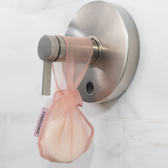The Soap Bar Bag by Kitsch, labeled Conditioner, hangs from your metallic shower faucet handle. Its pink mesh design integrates seamlessly into your routine against the white tiled wall, allowing optimal airflow for quick drying.