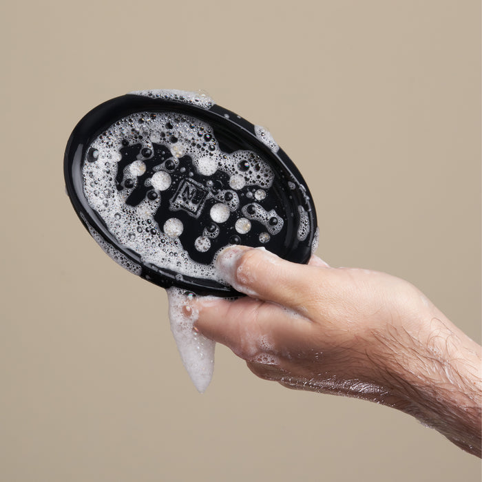 A hand holds the sleek NOT!CE Hair Co Bar Dish, adorned with soap suds, against a beige background.