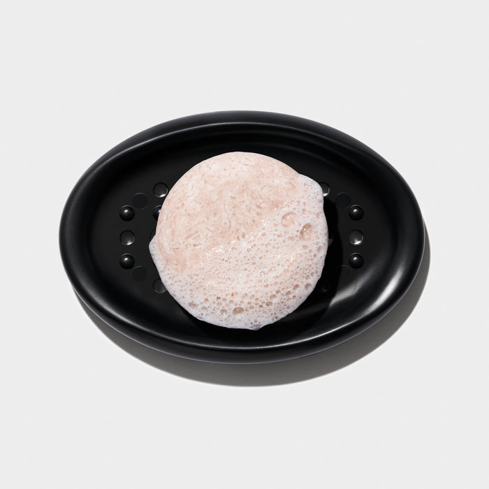 A light pink soap bar with frothy bubbles sits on a sleek NOT!CE Hair Co Bar Dish with drainage holes, against a white background, offering an elegant plastic-free hair care solution.