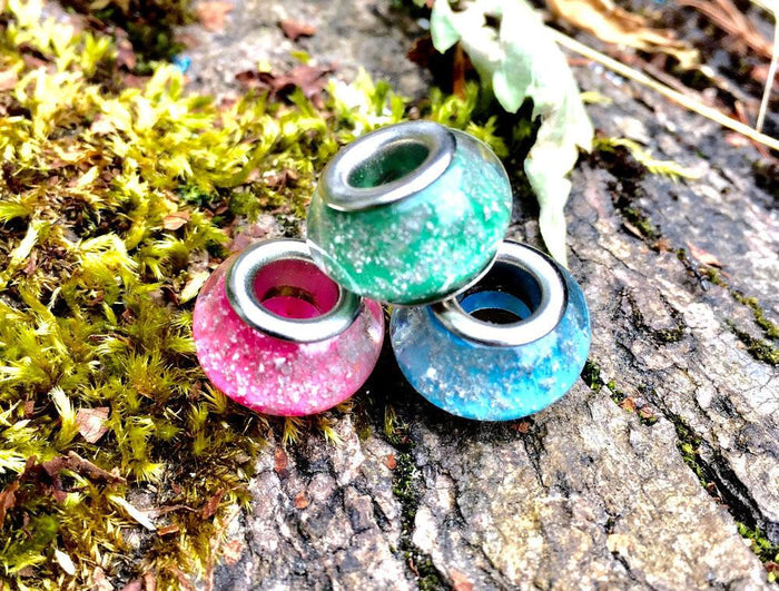 Three speckled cremains beads pink green blue with metal inner cores on a bark surface.