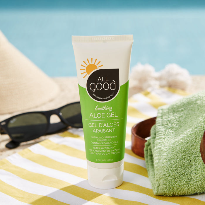 An Ultra Moisturizing Aloe Gel by All Good sits on a beach towel next to sunglasses and a green cloth, with a blurred beach and ocean backdrop creating a summery, coastal vibe.
