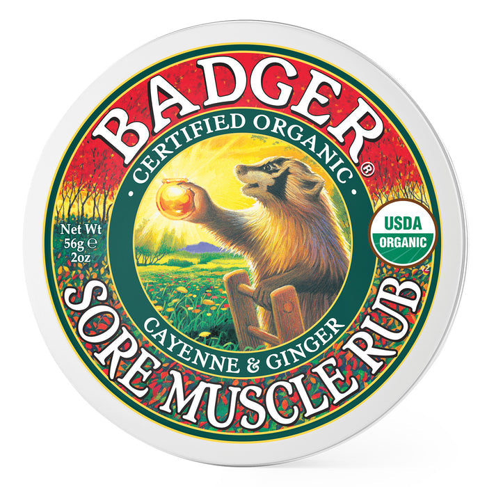 Round tin labeled Badger Balm Sore Muscle Rub, adorned with a badger and an orange in a field. It reads, Certified Organic, Cayenne & Ginger, USDA Organic, and Net Wt 56g 2oz. This warming balm is ideal for soothing sore muscles, set against a green and red design.
