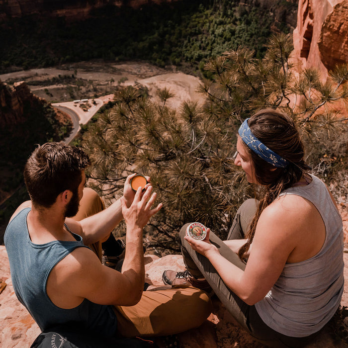 A man and woman sit on a rocky ledge overlooking a valley. The man holds Badger Balm’s Sore Muscle Rub, while the woman holds a similar organic tin. They are dressed casually, with the woman wearing a headband. Pine trees and a winding road can be seen below.