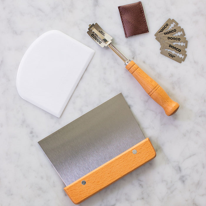 A marble surface displays the FarmSteady Sourdough Bread Making Kit with a wooden-handled dough scraper, white plastic scraper, DORCO-marked razor blades, a small brown leather pouch, and a wooden-handled razor tool—ideal for specialty baking equipment lovers.
