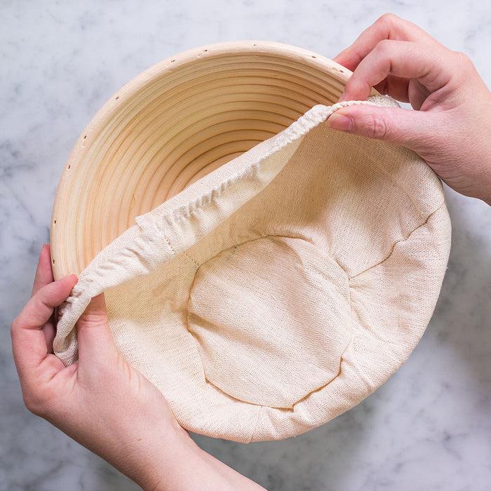 Two hands hold a FarmSteady Sourdough Bread Making Kits round banneton basket lined with natural fabric. Set on marble, one hand lifts the liners edge, offering a peek into essential specialty baking equipment.