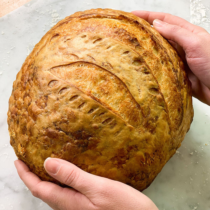 Two hands cradle a golden-brown, intricately scored sourdough loaf on a floured surface, highlighting its rustic appeal. Crafted with perfection using the FarmSteady Sourdough Bread Making Kit.