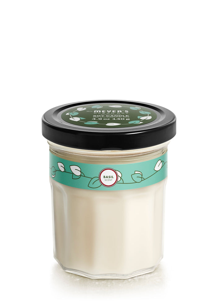 The Mrs. Meyers Soy Glass Jar Candle features a black-lidded glass jar with white cream, adorned with a green label and white leaves. The label reads Mrs. Meyers Clean Day and includes a Basil circle indicating its cruelty-free vegetable soy wax blend.