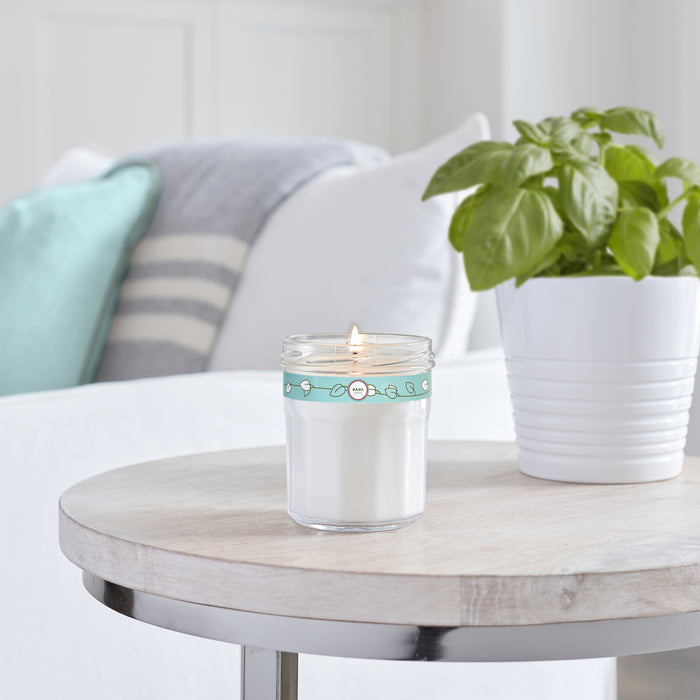 A Soy Glass Jar Candle by Mrs. Meyers glows warmly on a wooden table beside a white pot with a green plant. In the background, a white couch is adorned with a gray and white striped blanket and light blue pillow in the softly lit, cruelty-free space.