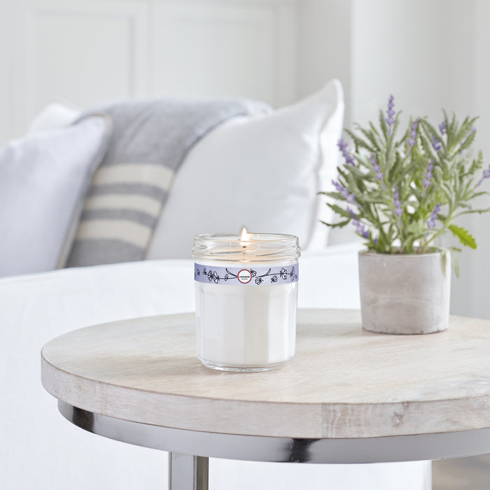 A Mrs. Meyers Soy Glass Jar Candle with a blue band sits lit on a wooden table next to a gray lavender pot. A white sofa with gray and white cushions is in the background, creating a cozy and cruelty-free atmosphere.