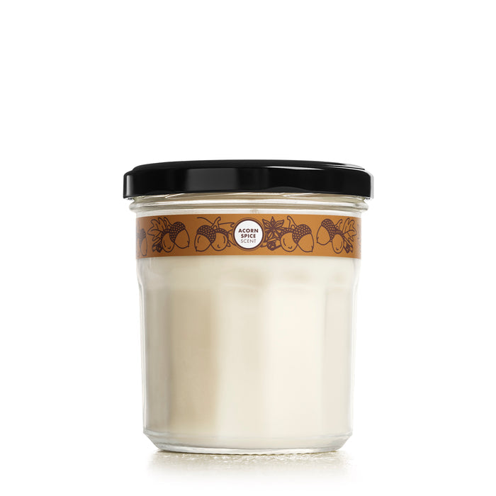 The Mrs. Meyers Soy Glass Jar Candle is a glass jar with a black lid and contains a white substance. It features a decorative brown label with acorns and the words ACORN and CREME, emphasizing its cruelty-free, vegetable soy wax composition.