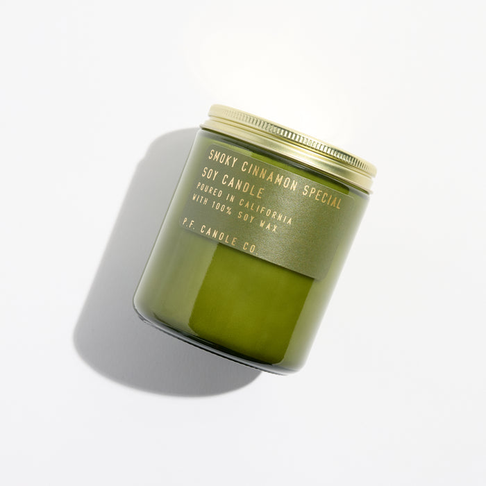 The Smoky Cinnamon Special Soy Candle in a green jar with a gold lid by P.F. Candle Co. casts an elegant shadow on white, labeled Poured in California with 100% Soy Wax. This Amber Jar exudes fresh, earthy vibes, perfect for any space.