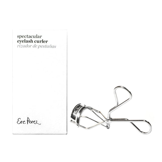A silver Spectacular Eyelash Curler by Ere Perez is elegantly displayed next to its white box, which says spectacular natural curl in Spanish. The box includes a signature-style logo and highlights hypoallergenic silicone pads for safe, stunning curls.