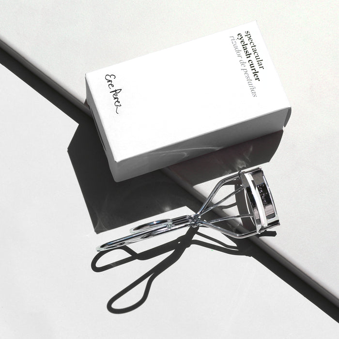 The Ere Perez Spectacular Eyelash Curler, featuring hypoallergenic silicone pads, rests on a light gray surface beside its white box. The shadows of both items extend diagonally in the bright light, creating a minimalist and natural aesthetic.