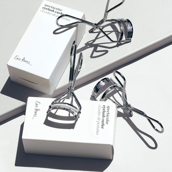 Three metallic Spectacular Eyelash Curlers by Ere Perez rest on a white surface, casting shadows that highlight their shiny texture. Equipped with hypoallergenic silicone pads, they offer a gentle touch for beautifully natural curls.
