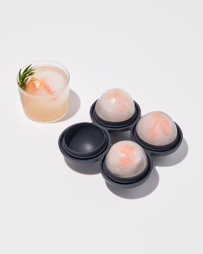 A set of three seamless ice spheres made with the W&P Sphere Ice Tray, each containing a slice of orange, sits beside a glass of pink drink. The drink is garnished with rosemary and features its own slow-melting sphere with an orange slice inside.