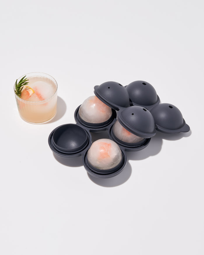 A glass of pink cocktail with a rosemary sprig sits beside W&Ps Sphere Ice Tray, featuring slow-melting ice spheres. The dark gray molds with transparent tops complement the vibrant drink against a sleek white background.