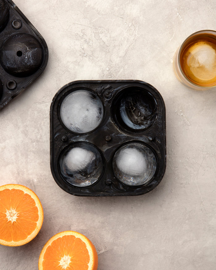 A W&P Sphere Ice Tray, in black, has four molds for large ice spheres. On a light textured surface, two of these slow-melting spheres are filled, while a glass nearby holds one alongside two fresh orange slices.