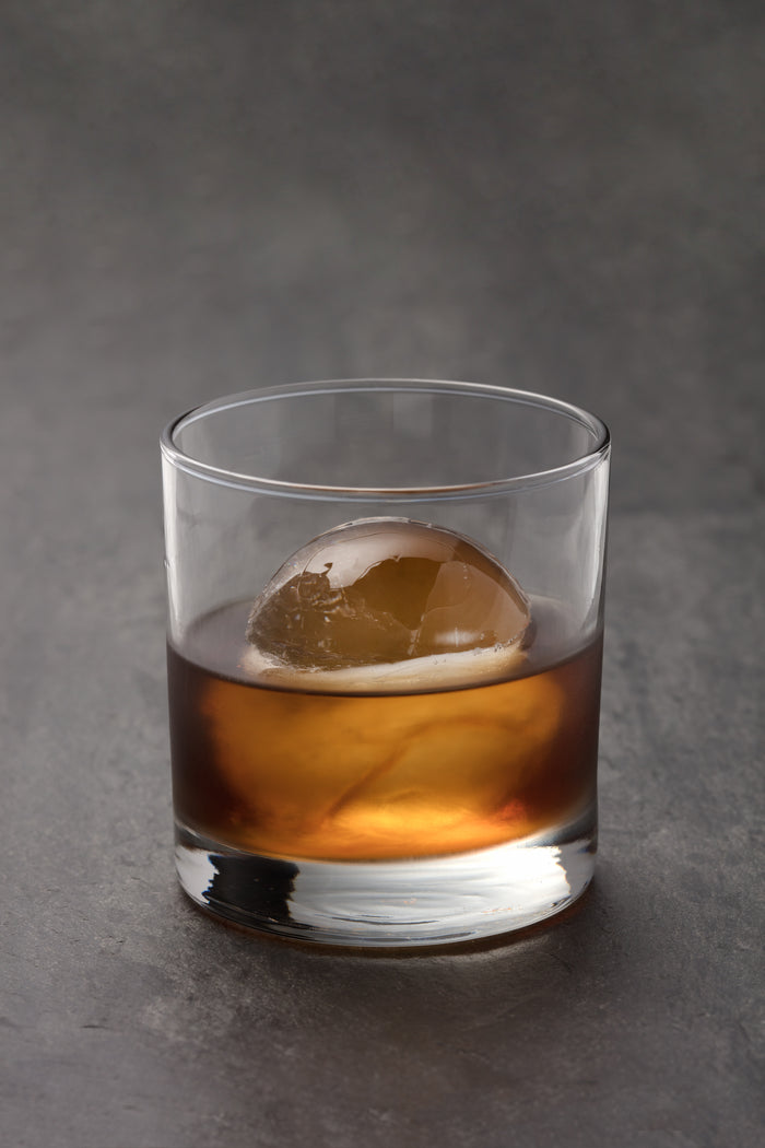 A glass of whiskey with a large ice sphere from the W&P Sphere Ice Tray rests on a dark, textured surface. The amber liquid contrasts with the clear, seamless sphere, giving it an elegant and sophisticated look.