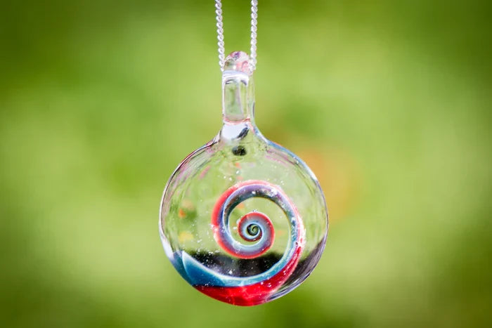 Close up of a colorful glass spiral pendant infused with ashes, hung on a silver chain with a bright green outdoor backdrop