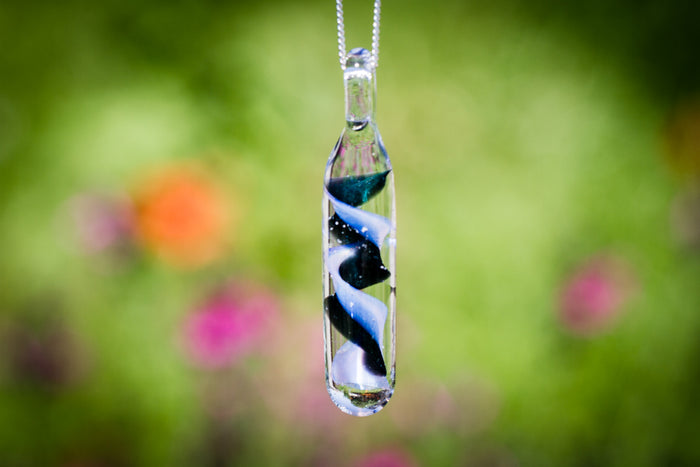Spiral blue black and white glass cylinder pendant on a silver chain against a blurred garden background.