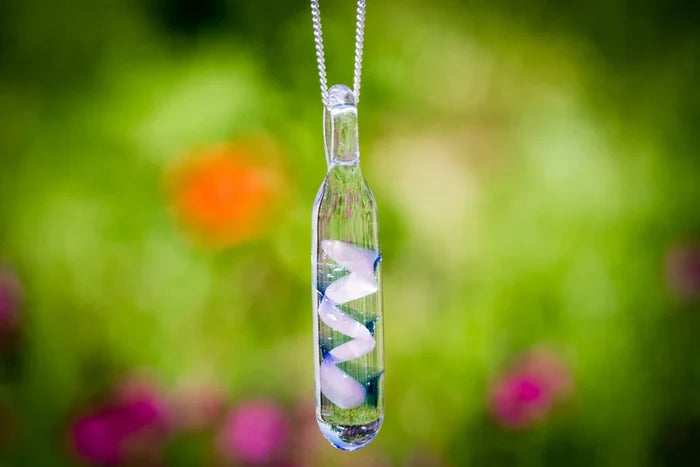 Clear glass spiral cylinder pendant with a white spiral inside, suspended from a silver chain against a blurred garden background