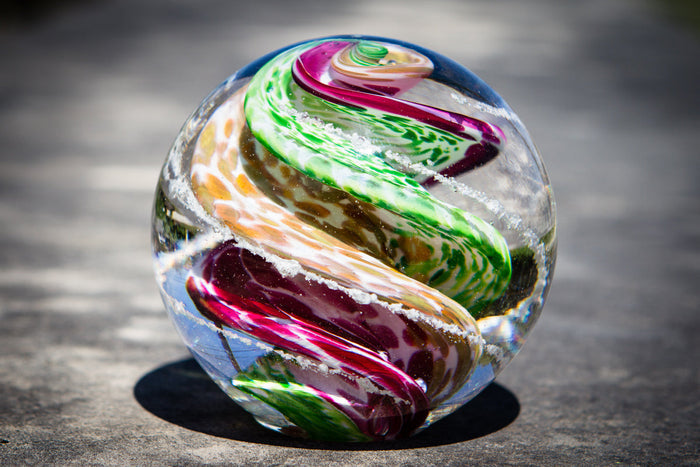 Swirled multicolor glass cremation orb with ashes infused, resting on a flat-bottom outdoor stone surface.