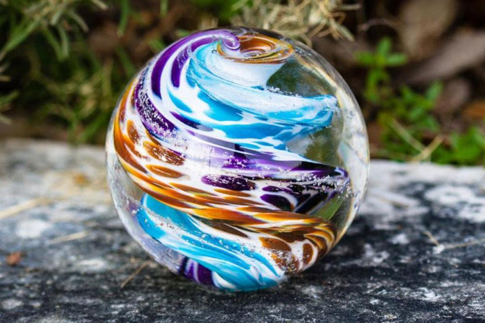Vibrant swirled glass cremation orb with blue orange and purple bands resting on a rough outdoor stone surface.