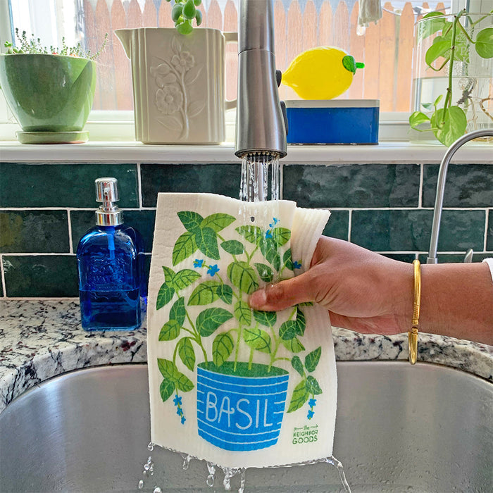 Someone washes a Sponge Cloth from The Neighborgoods, featuring a basil plant design, in the kitchen sink. Nearby, green and blue liquid soap dispensers flank the faucet as potted plants and fresh herbs enjoy sunlight on the windowsill above.