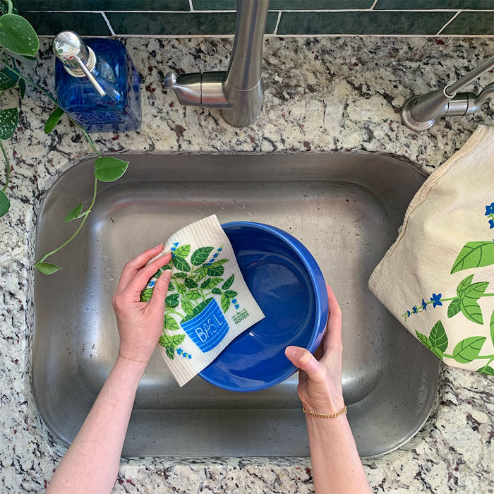 A person washes a blue bowl in a stainless steel sink using The Neighborgoods Sponge Cloth adorned with fresh herbs. The sink is set in a granite countertop, next to a green plant and blue bottle.
