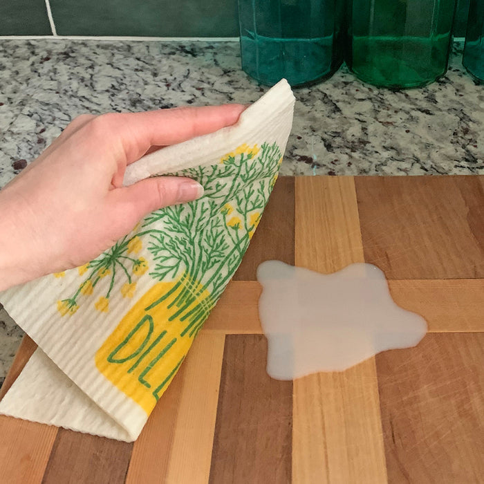A hand uses The Neighborgoods Sponge Cloth, featuring a green and yellow floral design, to wipe a spill on a wooden countertop while three green glass containers with fresh herbs sit in the background.