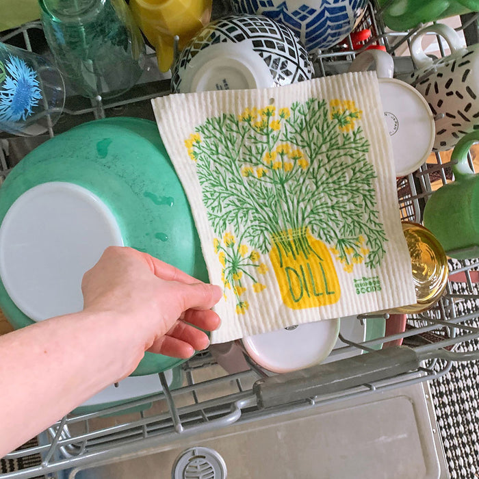 A hand places The Neighborgoods eco-friendly Sponge Cloth, featuring a dill plant illustration with a yellow pot labeled DILL and green leaves and flowers, into a dishwasher filled with colorful plates and cups.