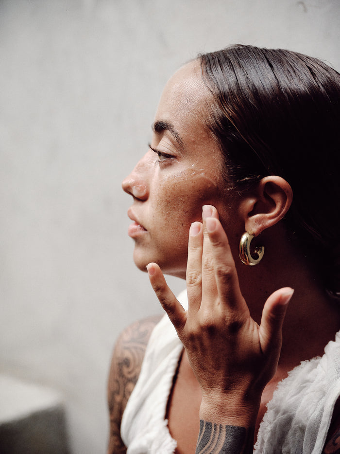 A person with a tattooed arm, in a white sleeveless top and gold hoop earrings, thoughtfully touches their cheek. Their hair is pulled back, showcasing naturally balanced skin in side profile against a light gray background. Achieve this glow with Thrive Natural Cares Spot-On Tone Balancing Serum.