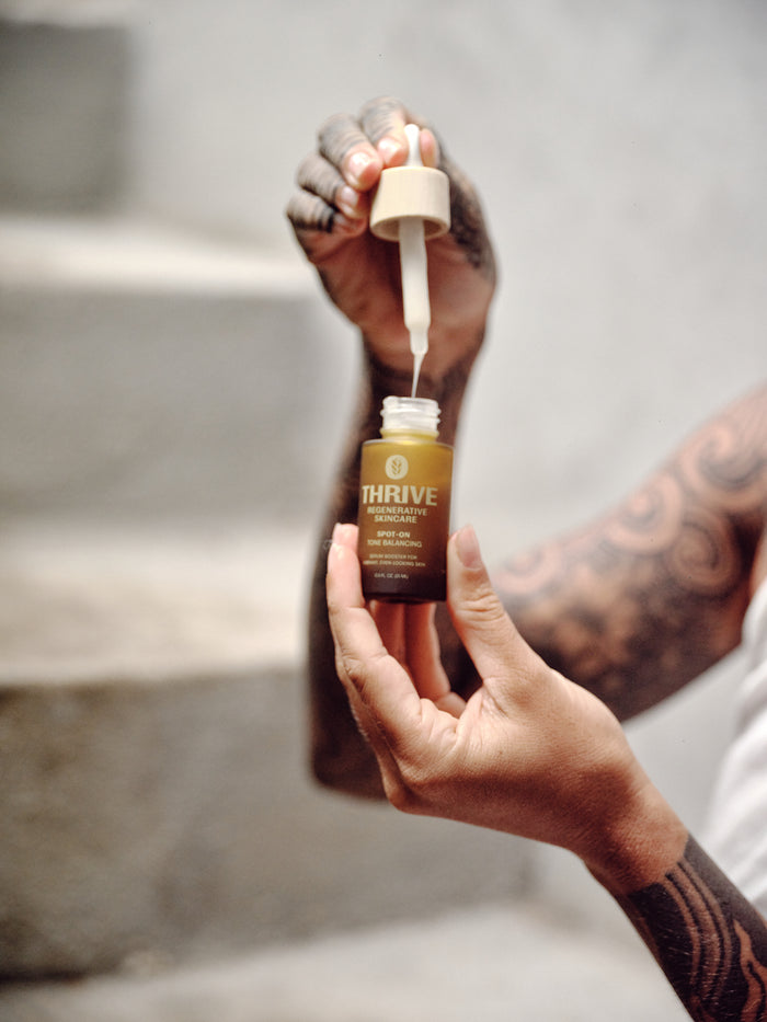 A person with tattooed arms holds a dropper above an open bottle labeled Spot-On Tone Balancing Serum by Thrive Natural Care. The indoor scene, featuring stone steps and a neutral background, shows them preparing to address dark spots and embrace their natural skin tone.