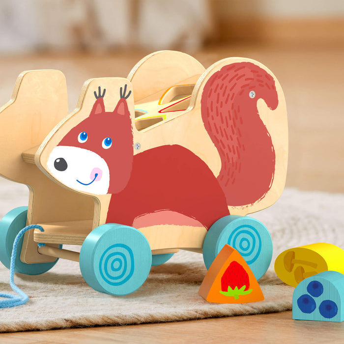 An eco-friendly Squirrel Pull Along Toy Sorting Box by HABA, featuring sustainably sourced wood, sits among colorful geometric blocks on a wooden floor. Includes a strawberry-themed triangle and semi-circle with blue cheese art—ideal for nurturing developmental skills.