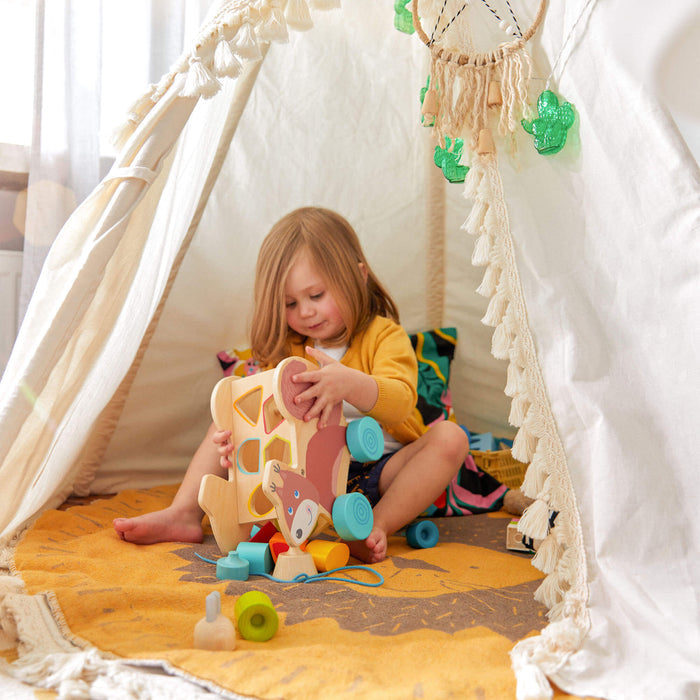 A child sits inside a white play tent, focusing on stacking colorful wooden blocks on a yellow patterned rug. With hanging decorations and tassels around, the childs skills blossom as they play in their yellow shirt with HABAs Squirrel Pull Along Toy Sorting Box.