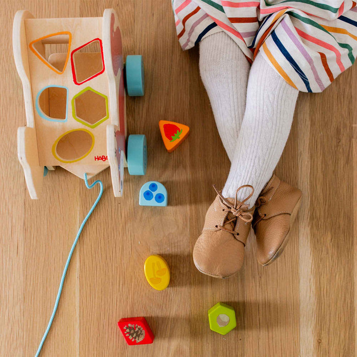 A child in a striped dress and white tights sits on a wooden floor, wearing tan shoes. Next to them, the HABA Squirrel Pull Along Toy Sorting Box with blue wheels aids skill development as colorful shapes are scattered around it.
